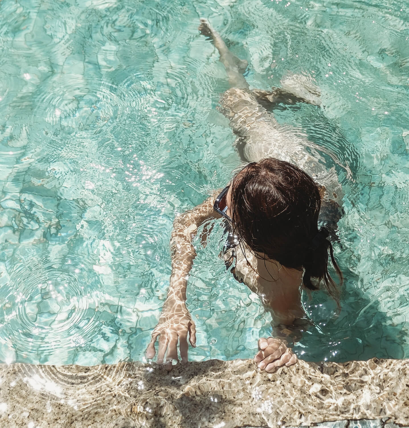 Overhead view of a person in a swimming pool with clear aqua water, touching the stone edge.