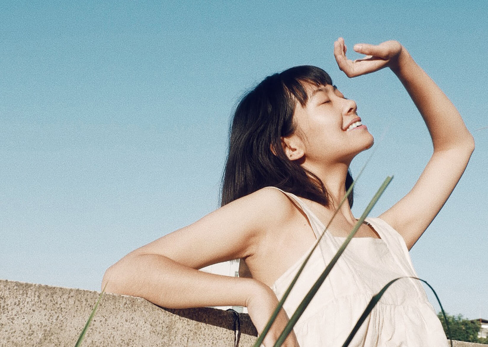 A person with dark hair and a white sleeveless top, smiling and leaning against a stone wall. Their hand is raised to shield their eyes from the sun.