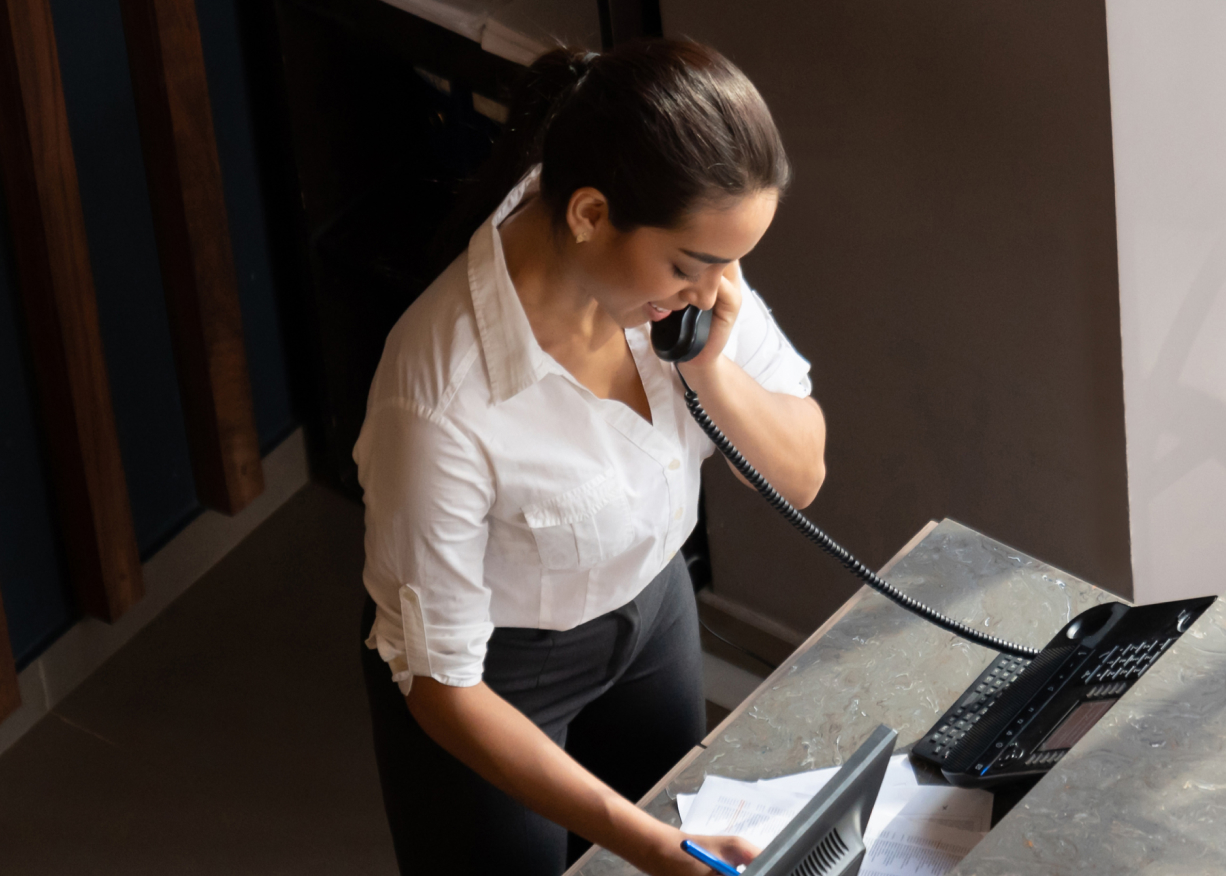 Overhead view of a person at a reception desk in a white collar shirt. They are on the phone and writing notes with their other hand.