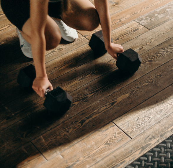 Close up of a person’s arms grabbing onto dumbbells resting on a timber floor.